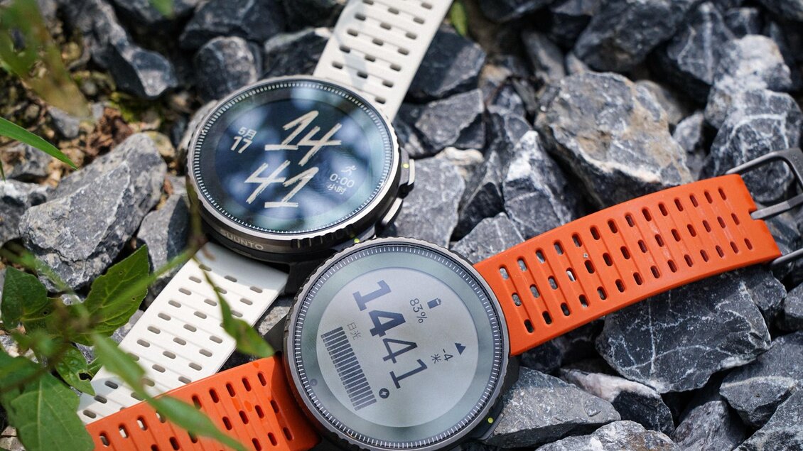 Zwei Sportuhren liegen auf einem Kiesboden. Eine hat ein weißes Armband, die andere ein oranges Armband. | © Suunto