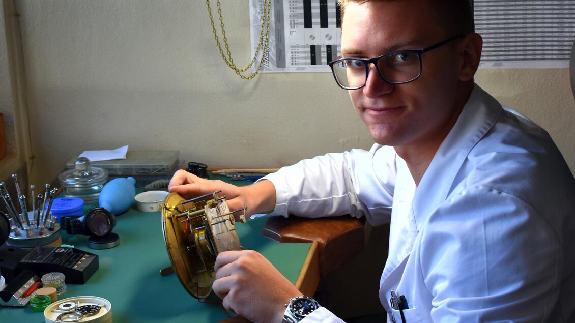Eine Person in einem weißen Laborkittel arbeitet an einem Uhrwerk. Der Arbeitsplatz ist gut ausgestattet mit Werkzeugen und Materialien. | © Heinz Michalka