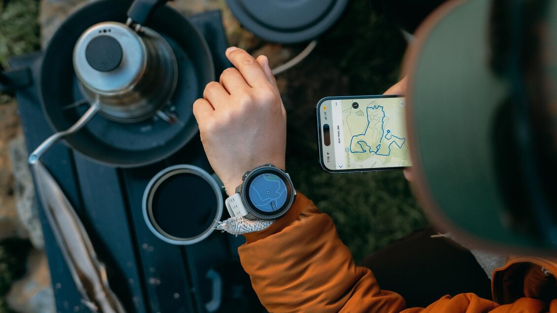 Eine Person sitzt im Freien und checkt eine smartwatch. Auf einem Handy wird eine Karte angezeigt, während ein Kaffeekocher im Hintergrund steht. | © Suunto