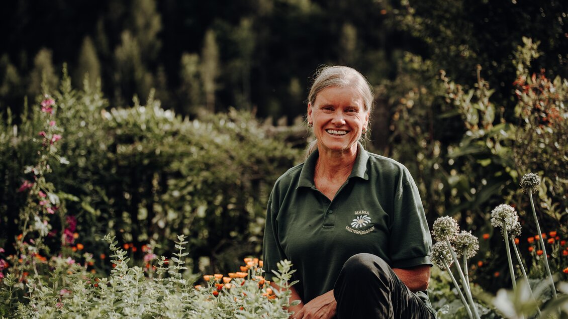 Eine lächelnde Frau sitzt in einem Garten voller Blumen und Pflanzen. Sie trägt ein grünes T-Shirt und wirkt glücklich in ihrer Umgebung. | © Freilichtmomente