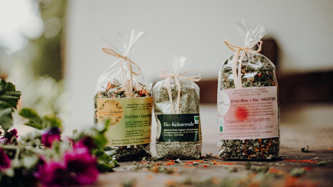 Drei transparent verpackte Kräuterpäckchen stehen auf einem Tisch. Im Hintergrund sind einige frische Blumen sichtbar. | © Freilichtmomente