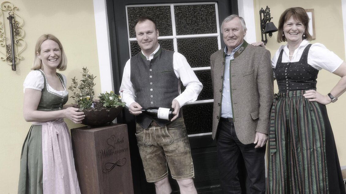 Eine Gruppe von vier Personen in traditioneller bayerischer Tracht steht vor einem Haus. Sie lächeln und präsentieren eine Flasche Sekt. | © www.michilorenz.at