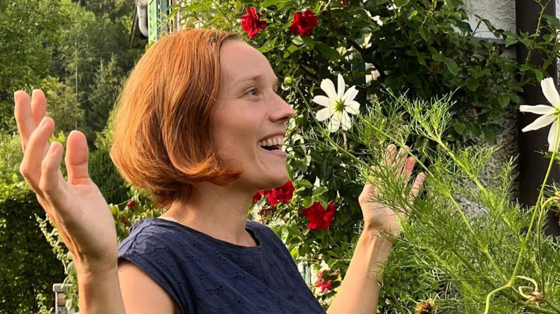 Eine lächelnde Frau mit kurzen, roten Haaren steht vor blühenden Pflanzen. Sie hebt die Hände und strahlt Freude aus. | © Birgit Bossert