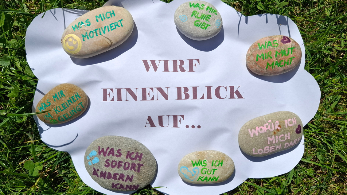 Eine Gruppe von Steinen mit bunten Texten liegt auf einem weißen Blatt im Gras. Die Texte beinhalten Fragen zur Selbstreflexion und Motivation. | © Birgit Bossert