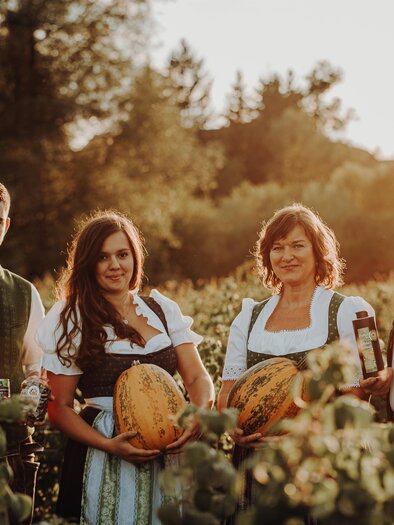 Eine Gruppe von vier Personen in traditioneller Kleidung steht in einem Weinberg bei Sonnenuntergang. Sie halten Melonen und Flaschen in den Händen. | © Freilichtmomente