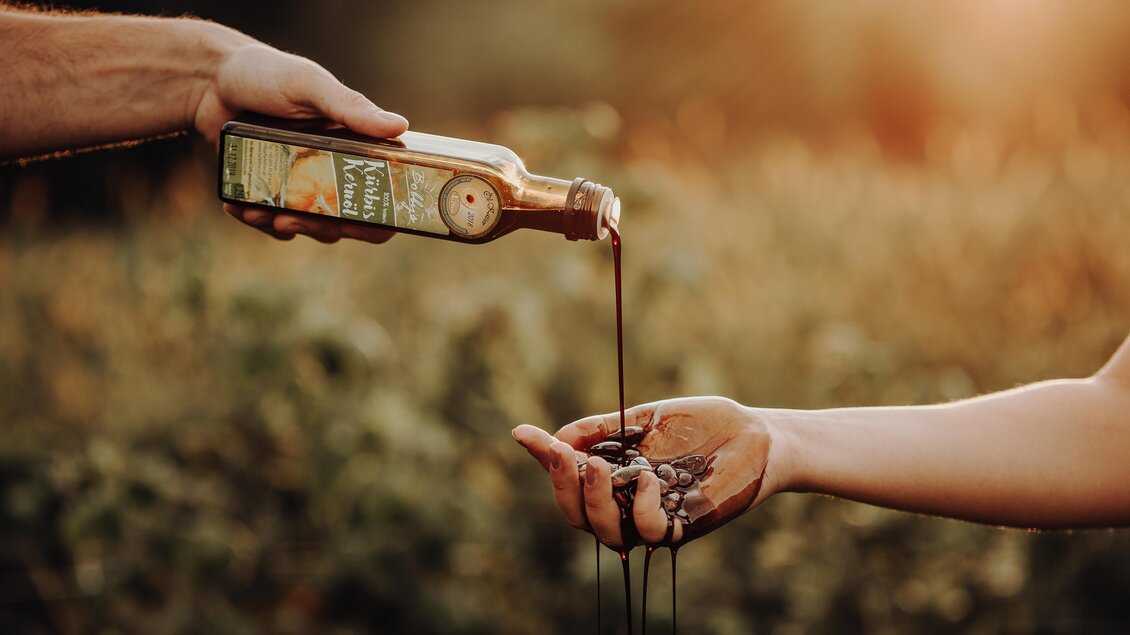 Eine handgefertigte Flasche wird über eine andere Hand gehalten, aus der Olivenöl langsam herausfließt. Im Hintergrund ist eine grüne Landschaft bei Sonnenuntergang zu sehen. | © Freilichtmomente