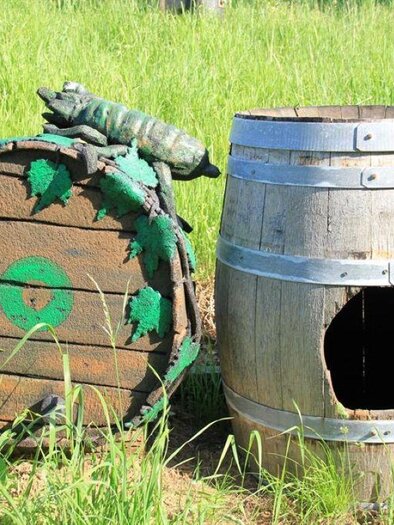 Ein bemaltes Holzfass und eine verzierte, künstlerisch gestaltete Kiste liegen im Gras. Auf der Kiste sitzt eine kleine, grüne Eidechse. | © KulturWeingut Kästenburg