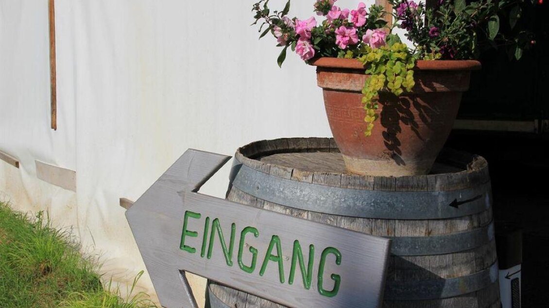 Ein Holzfass mit einem blühenden Topf, auf dem ein grünes Schild mit der Aufschrift „EINGANG“ steht. Dahinter ist ein Zelt mit einer hellen Stoffwand sichtbar. | © Ulrike Elsnegg