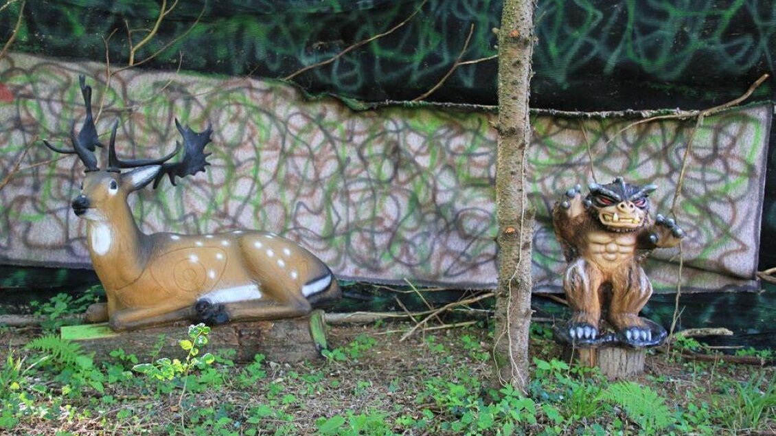 Zwei dekorative Figuren im Wald: Ein Hirsch auf der linken Seite und ein Monster auf der rechten Seite. Im Hintergrund sieht man eine bemalte Wand mit bunten Mustern. | © Ulrike Elsnegg