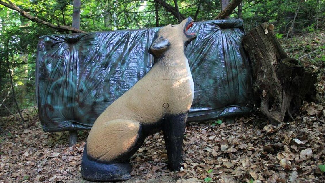 Eine Kunstfigur eines Tieres sitzt im Wald. Im Hintergrund ist eine schwarze Plane und trockene Blätter zu sehen. | © Ulrike Elsnegg