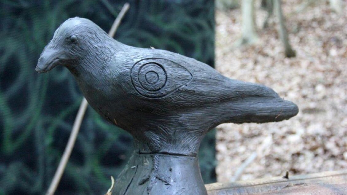 Eine schwarze Vogelstatue steht auf einem Holzstamm im Wald. Der Hintergrund besteht aus verschwommenen Baumstämmen und Laub. | © Ulrike Elsnegg