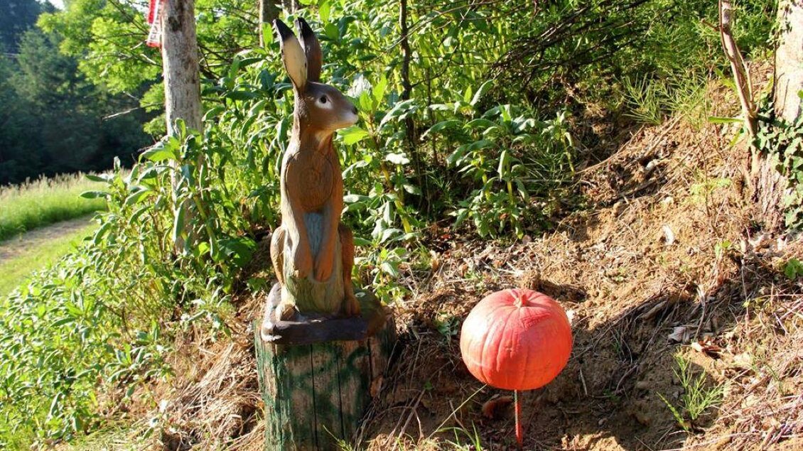 Eine geschnitzte Holzfigur eines Hasen sitzt auf einem Baumstumpf. Daneben liegt ein orangefarbener Ballon auf dem Boden. | © Ulrike Elsnegg