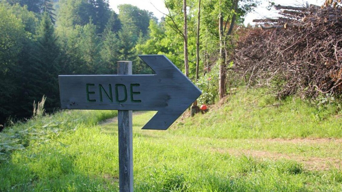 Ein Schild mit der Aufschrift „ENDE“ zeigt nach rechts. Im Hintergrund ist eine grüne Landschaft mit Bäumen und Holzstapel zu sehen. | © Ulrike Elsnegg