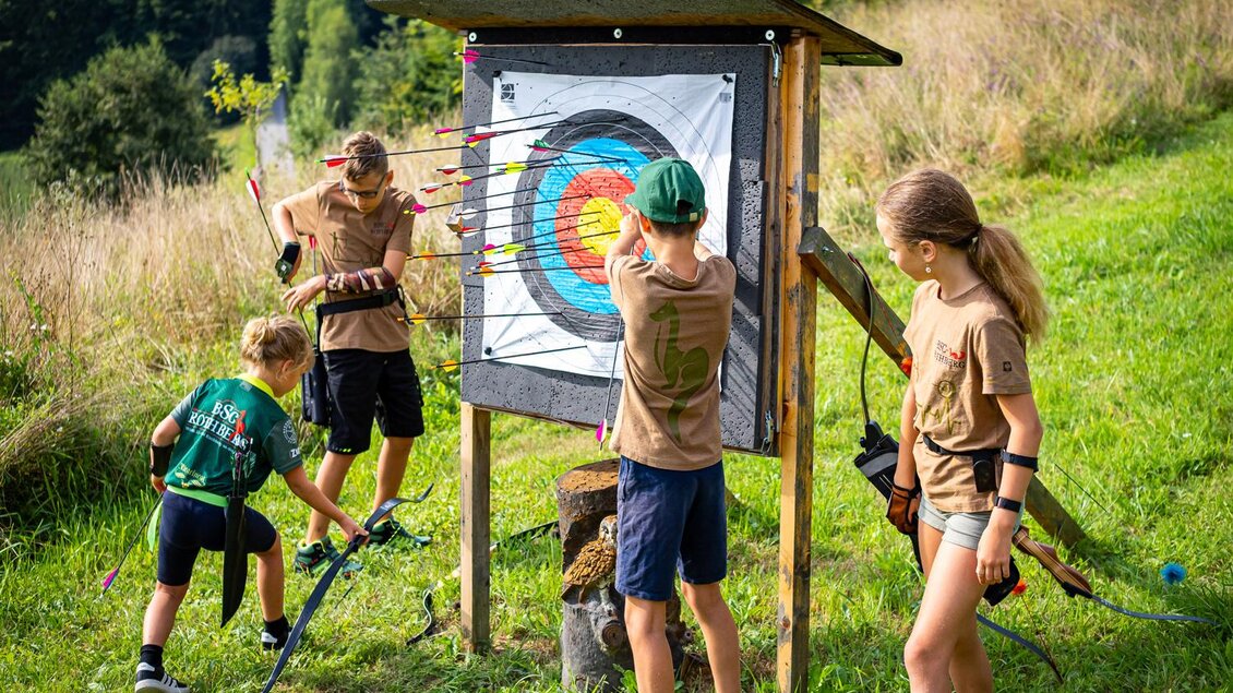 Kinder üben das Bogenschießen auf einem Übungsplatz im Freien. Im Hintergrund sind Bäume und eine grüne Wiese sichtbar. | © Andreas Schlögl