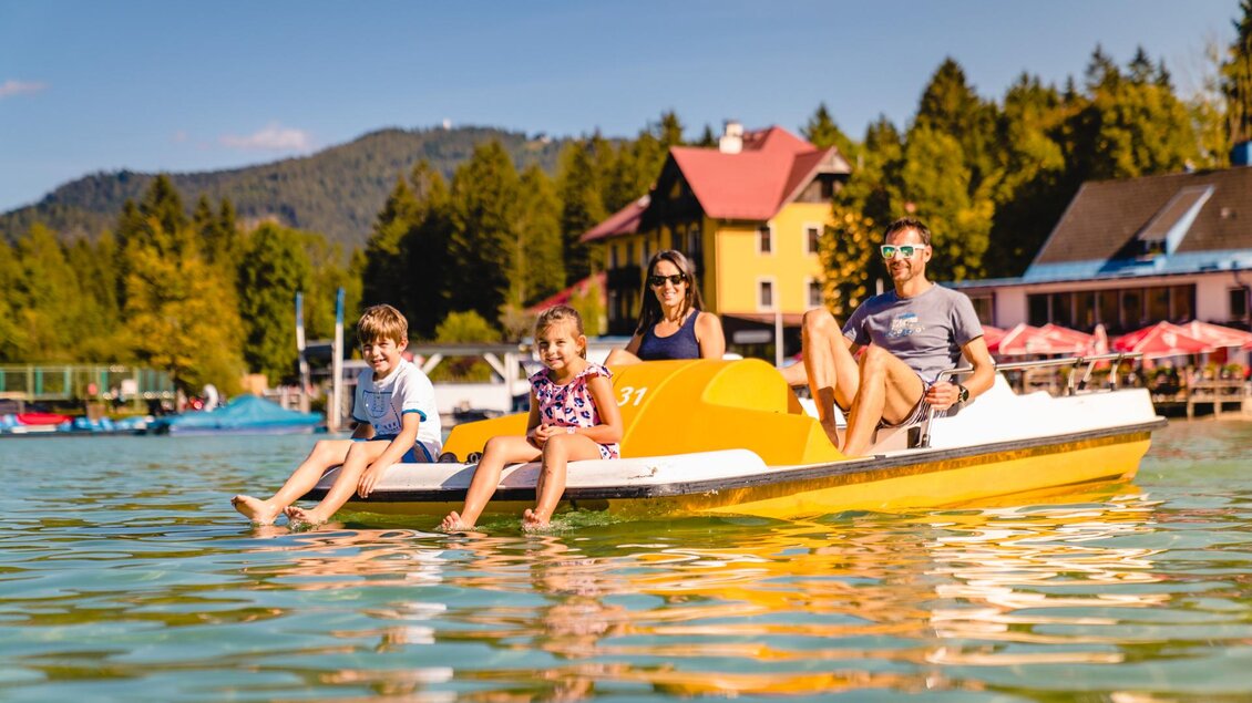Eine Familie genießt eine Bootsfahrt auf einem ruhigen See. Im Hintergrund sind Bäume und farbige Gebäude zu sehen. | © TV Hochsteiermark