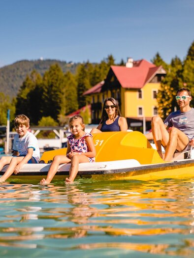 Eine Familie genießt eine Bootsfahrt auf einem ruhigen See. Im Hintergrund sind Bäume und farbige Gebäude zu sehen. | © TV Hochsteiermark