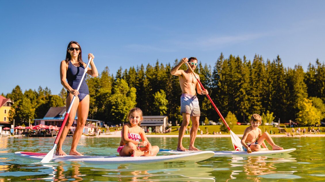 Eine Familie paddelt auf dem Wasser mit Stand-Up-Paddle-Boards. Im Hintergrund sind Bäume und eine sonnige Landschaft zu sehen. | © TV Hochsteiermark