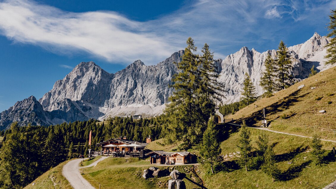 Eine malerische Alpenlandschaft mit hohen Bergen im Hintergrund. Im Vordergrund sieht man traditionelle Holzhäuser, umgeben von grünem Gras und Bäumen. | © Christine Höflehner