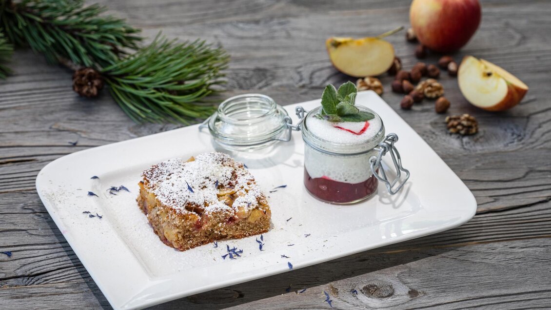Ein köstliches Dessert auf einem weißen Teller. Es besteht aus einem Stück Kuchen mit Puderzucker und einem kleinen Glas mit einer Joghurtspeise, garniert mit frischer Minze. | © Martin Huber