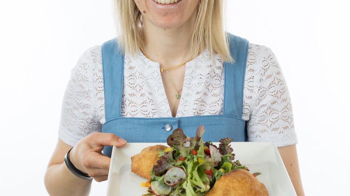 Eine lächelnde Frau in traditioneller Tracht hält einen Teller mit einem Salat und frittierten Speisen. Der Hintergrund ist schlicht und weiß.
