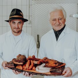Zwei Männer in weißen Schürzen halten ein Tablett mit verschiedenen Wurstwaren. Im Hintergrund ist eine metallene Arbeitsfläche zu sehen. | © GenussReich