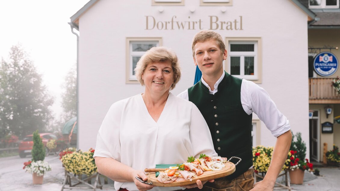 Ein freundliches Paar steht vor dem Restaurant Dorfwirt Bratl und hält ein Tablett mit Essen. Die Umgebung ist blühend und einladend. | © Dorfwirt Bratl