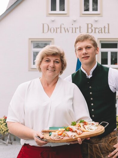 A friendly couple is standing in front of the restaurant Dorfwirt Bratl, holding a tray of food. The surroundings are blooming and inviting. | © Dorfwirt Bratl