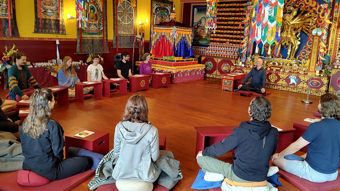 Eine Gruppe von Menschen sitzt in einem bunten Raum und hört einer Person zu, die spricht. Im Hintergrund sind verzierte Wandteppiche und spirituelle Symbole sichtbar. | © Dzokden Kalapa