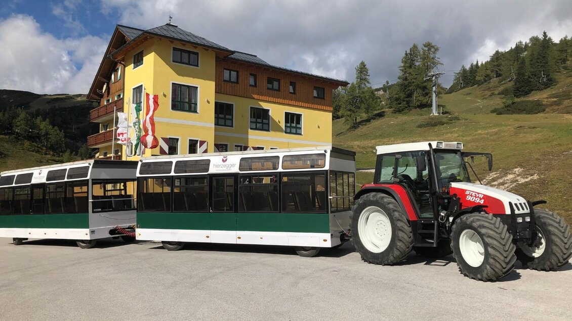 Ein Traktor zieht eine Bahn neben einem großen gelben Gebäude. Die Umgebung ist bergig mit Bäumen im Hintergrund. | © Hotel Hierzegger