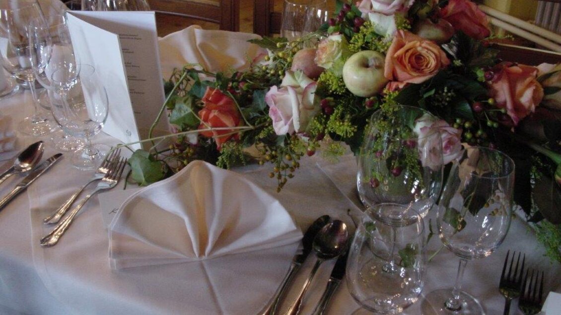 Ein elegant gedeckter Tisch mit Gläsern, Besteck und einer dekorativen Blumengesteck. Die Tischdecke ist weiß und ein Menü ist vorbereitet. | © Burg Deutschlandsberg