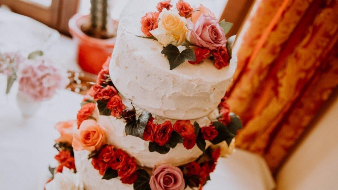 Eine elegante Torte mit mehreren Schichten, geschmückt mit roten und rosa Rosen. Im Hintergrund befinden sich einige Pflanzen und ein warmes Licht. | © Burg Deutschlandsberg
