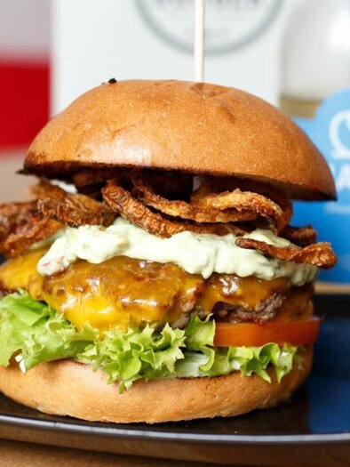 Ein köstlicher Burger mit geschmolzenem Käse, frischem Gemüse und knusprigen Zwiebelringen. Im Hintergrund sieht man ein Getränk und ein elegantes Glas. | © Burger Factory