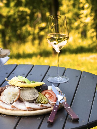 Ein appetitliches Brett mit frischen Lebensmitteln, Brot und Schinken liegt auf einem Tisch. Im Hintergrund steht ein Glas Weißwein vor einer grünen Wiese. | © TVB Thermen- und Vulkanland