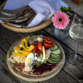 Ein buntes Tellergericht mit verschiedenen Wurst- und Käsesorten, frischem Gemüse und einem gekochten Ei. Neben dem Teller steht ein Glas Weißwein und eine dekorative Blume. | © Tom Lamm