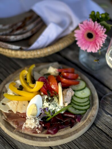 Ein buntes Tellergericht mit verschiedenen Wurst- und Käsesorten, frischem Gemüse und einem gekochten Ei. Neben dem Teller steht ein Glas Weißwein und eine dekorative Blume. | © Tom Lamm