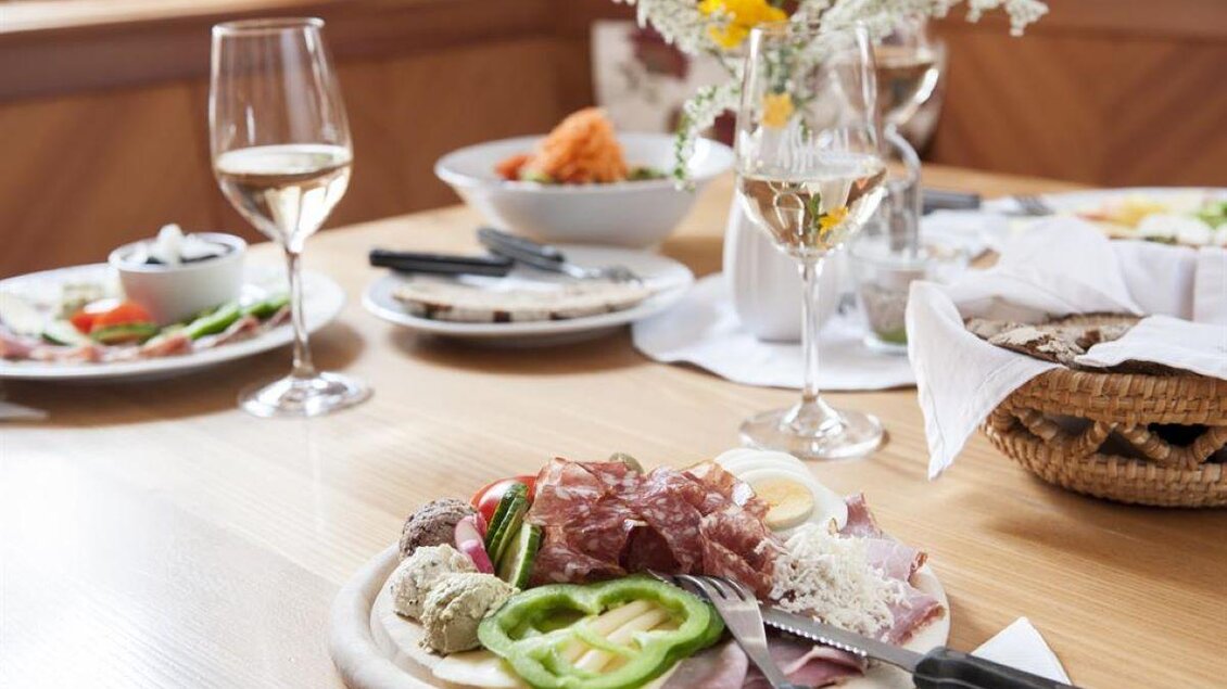 Ein schön gedeckter Tisch mit einer Auswahl an Aufschnitt, Gemüse und einem Glas Wein. Im Hintergrund sind weitere Gerichte und eine Blumenvase zu sehen. | © Weingut Assigal
