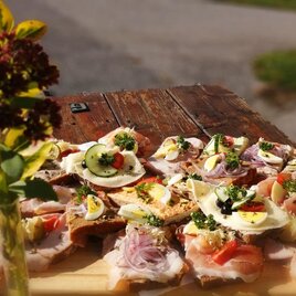 Ein Holzbrett mit verschiedenen belegten Broten, garniert mit frischem Gemüse und Eiern. Im Hintergrund steht eine Blume, die einen Hauch von Natur hinzufügt. | © Weingut Buschenschank Bernhard