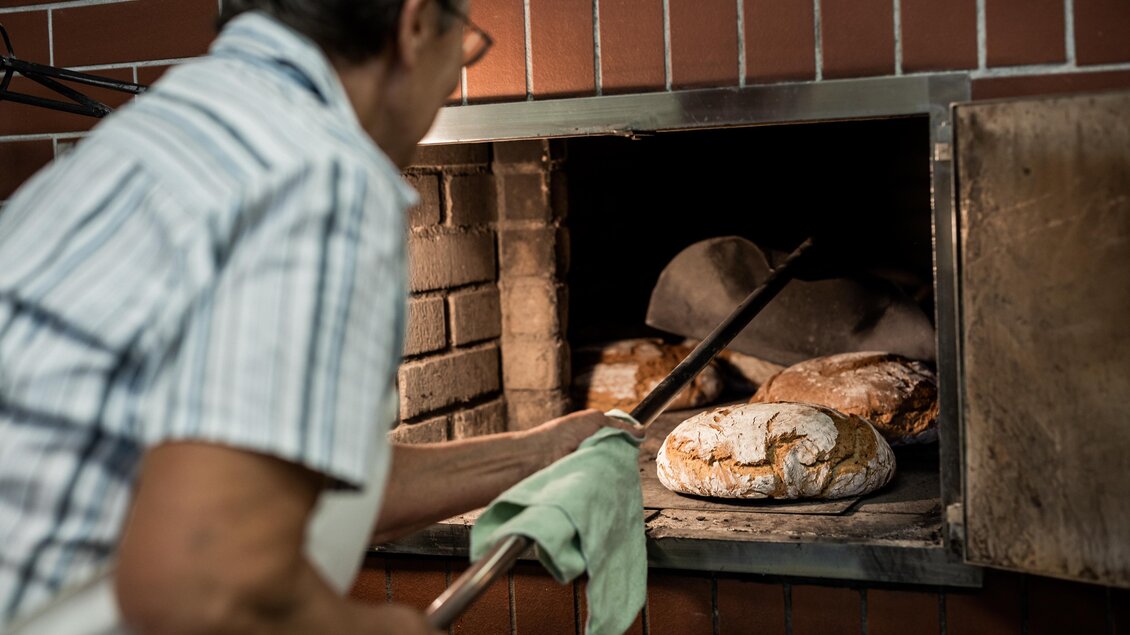 Ein Bäcker holt frisch gebackenes Brot aus dem Ofen. Im Hintergrund sind weitere Brote sichtbar. | © BROBOTERS