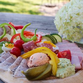 Ein gefülltes Holbrett mit verschiedenen Wurst- und Käseaufschnitten, frischem Gemüse und Dip. Im Hintergrund steht eine weiße Blume. | © Weingut & Buschenschank Bockmoar