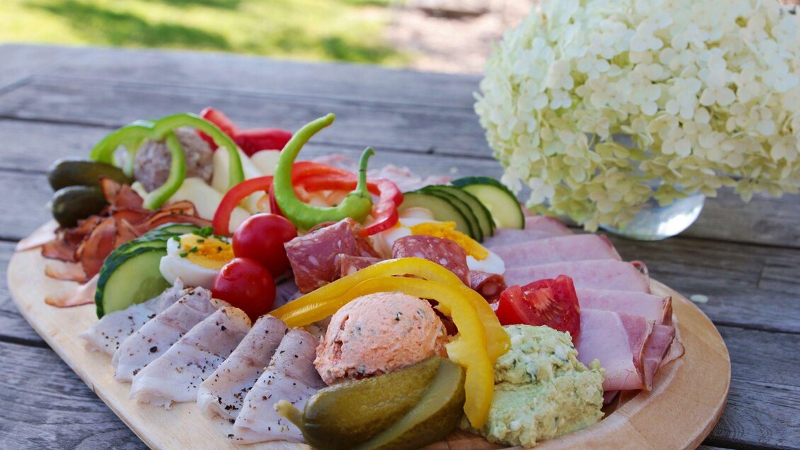 Ein gefülltes Holbrett mit verschiedenen Wurst- und Käseaufschnitten, frischem Gemüse und Dip. Im Hintergrund steht eine weiße Blume. | © Weingut & Buschenschank Bockmoar