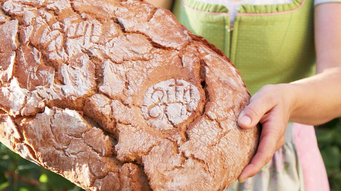 Eine Person hält ein großes, frisch gebackenes Brot in den Händen. Das Brot hat eine rustikale Kruste und ist mit Mustern verziert. | © Weingut & Buschenschank Bockmoar