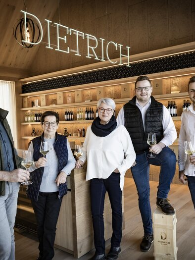 Eine Gruppe von fünf Personen steht in einem modernen Weingeschäft. Im Hintergrund sind Holzregale mit Weinflaschen und der Schriftzug "DIETRICH" zu sehen. | © BU Dietrich