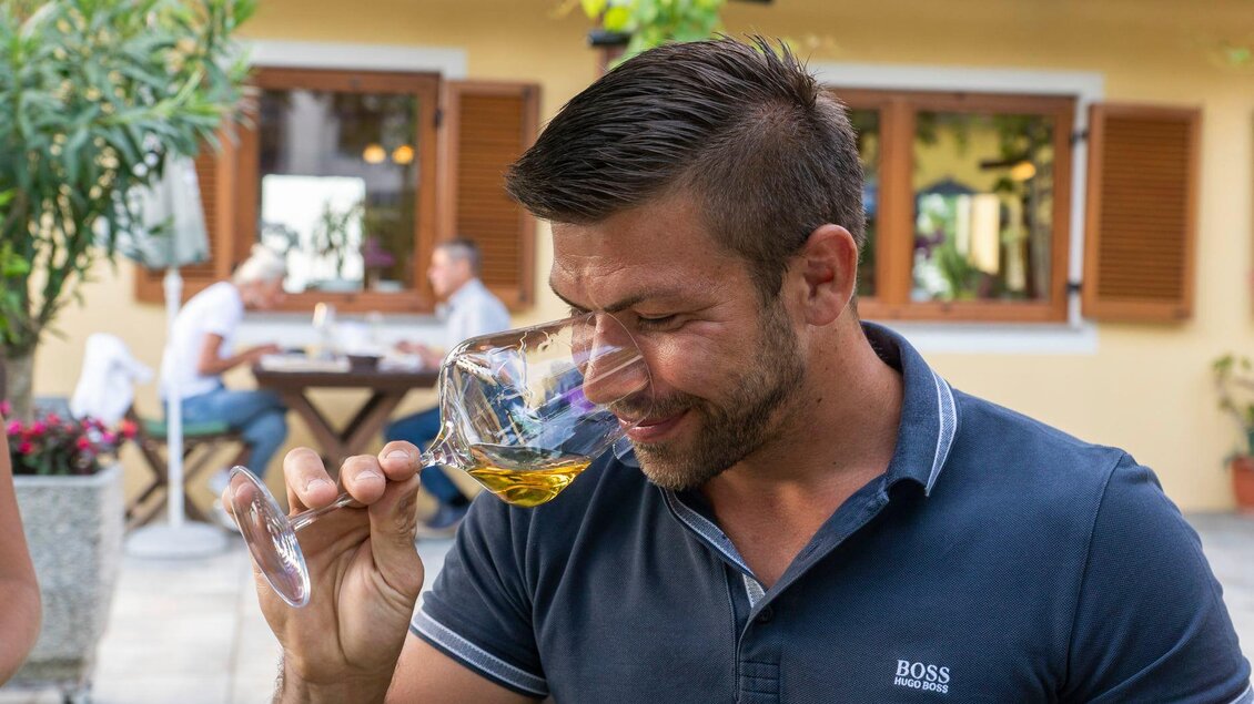 Ein Mann betrachtet aufmerksam ein Glas Wein. Im Hintergrund sind unscharf weitere Personen und eine gemütliche Außenatmosphäre zu sehen. | © Buschenschank & Weingut Gallunder