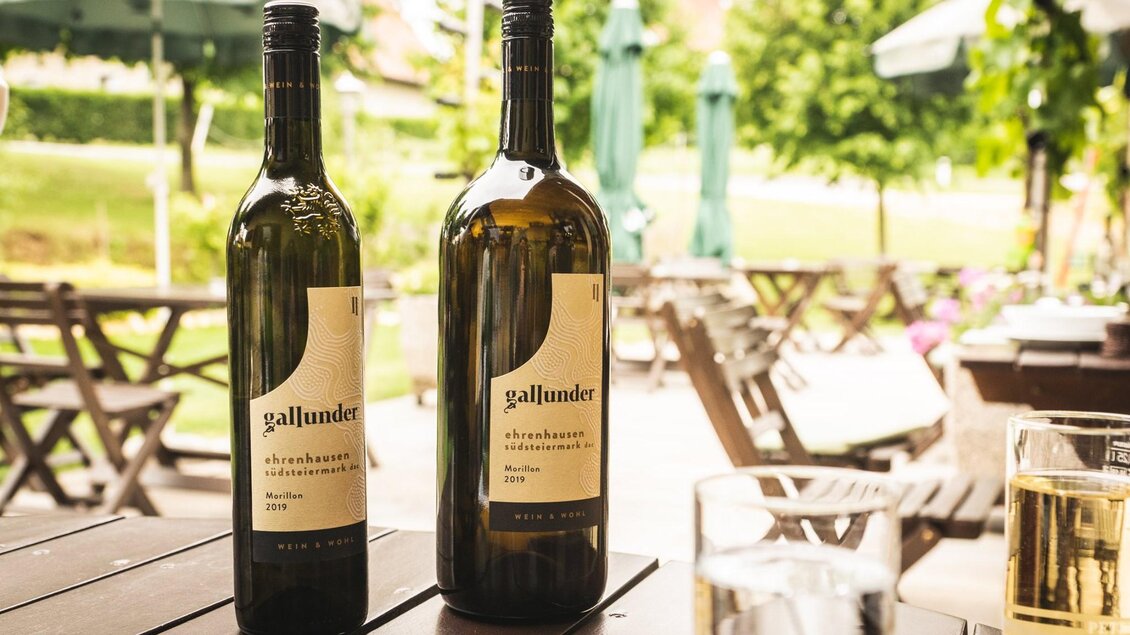 Zwei Weinflaschen stehen auf einem Tisch in einem sonnigen Garten. Im Hintergrund sind Sitzgelegenheiten und grüne Bäume zu sehen. | © Buschenschank & Weingut Gallunder
