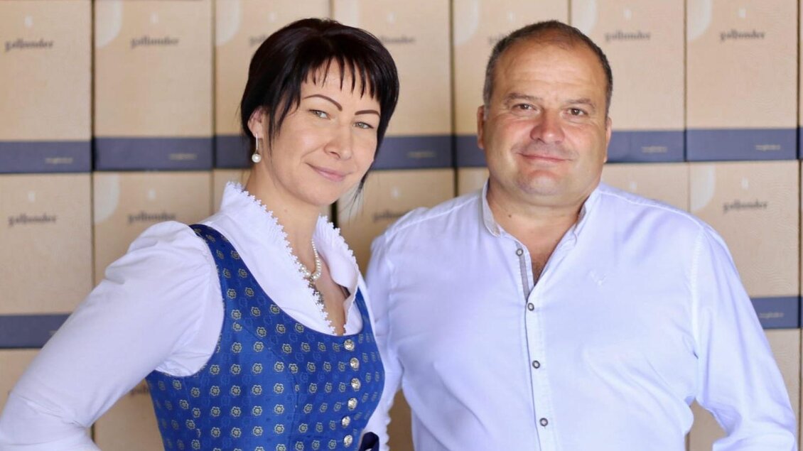 Ein Paar steht vor einer Wand mit kistenförmigen Verpackungen. Die Frau trägt ein traditionelles Kleid und der Mann ein weißes Hemd. | © Weingut & Buschenschank Gallunder