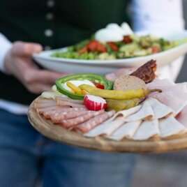 Ein Holzbrett mit verschiedenen Sorten Aufschnitt, darunter Schinken und Wurst, dekoriert mit Gemüse. Im Hintergrund hält jemand einen Salat. | © Michaela Lorber