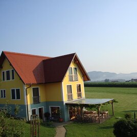 Ein charmantes gelbes Haus mit rotem Dach steht in einer grünen Landschaft. Im Hintergrund sind sanfte Hügel und ein klarer Himmel zu sehen. | © Winzerhof Grebenz