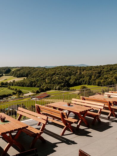 Eine sonnige Terrasse mit vielen Holztischen und Bänken. Im Hintergrund erstreckt sich eine grüne Landschaft unter einem klaren blauen Himmel. | © Familie Hartinger