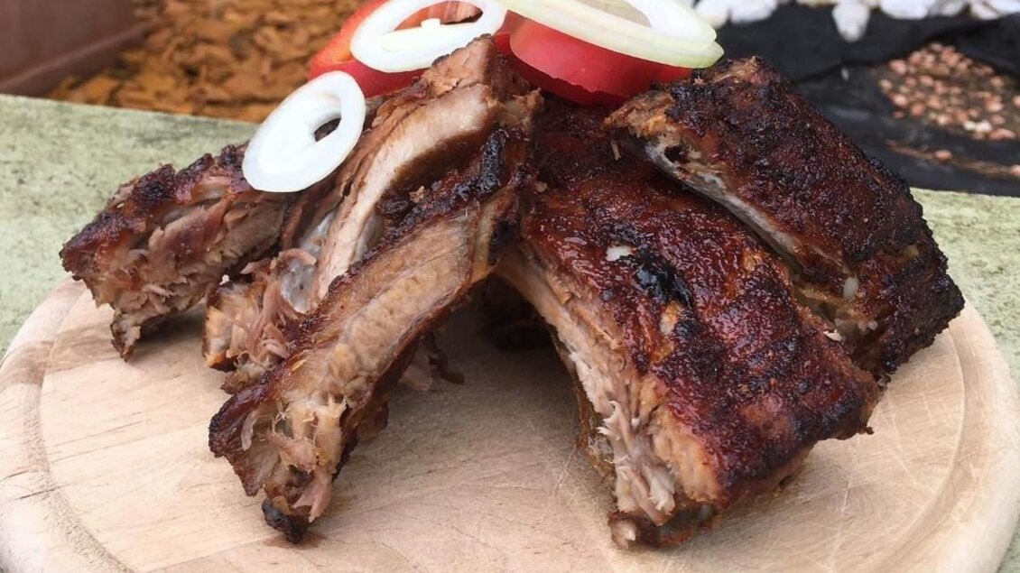 Saftige Rippchen auf einem Holzbrett, garniert mit Zwiebelringen und Tomatenscheiben. Eine appetitliche Präsentation für ein köstliches Gericht. | © Häuser am Wald | T. Hartner