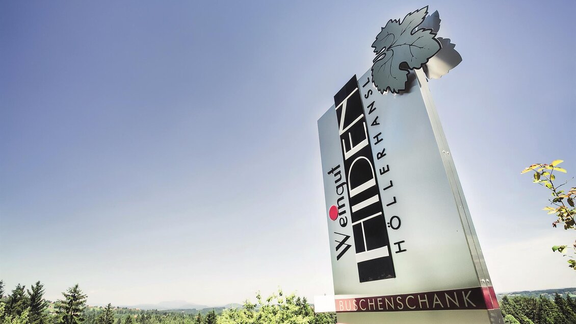 Ein modernes Schild mit dem Schriftzug „Höllermann“ und einem Weinblatt. Im Hintergrund sind grüne Bäume und ein klarer Himmel zu sehen. | © Schilcherland Steiermark